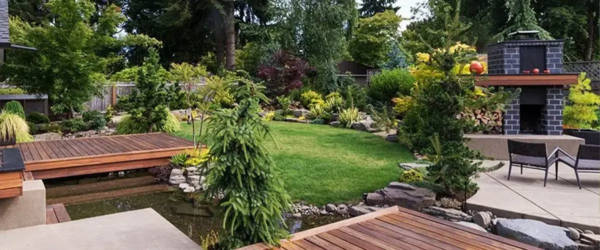 A large outdoor living space with an exterior oven made of dark bricks, two decks, a water feature, and a lot of shrubs, trees, and colored plants around