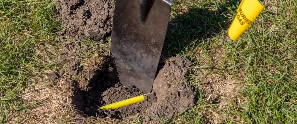 Buried plastic natural gas line in hole of yard. Shovel in soil and yellow buried gas warning flag marking location