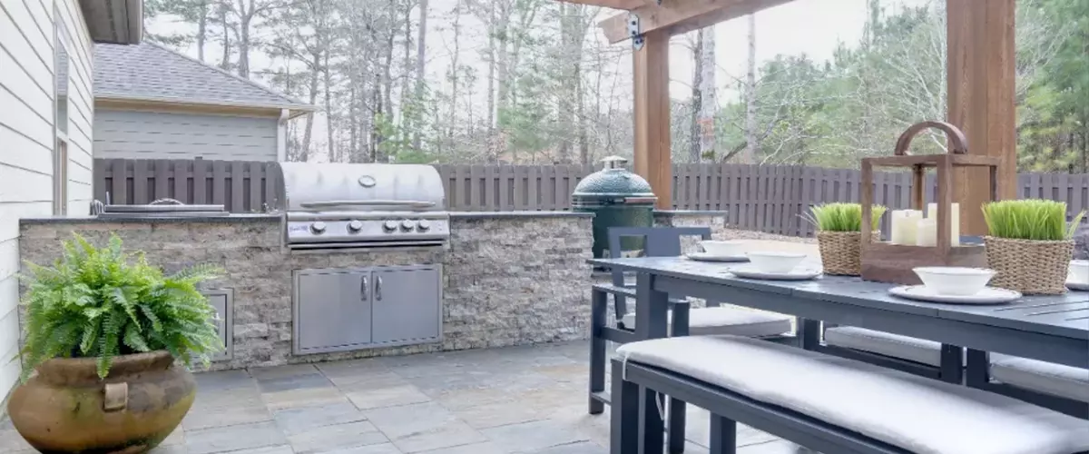 Pretty summer outdoor kitchen with table set and grilling station underneath wooden arbor on stone patio - Farrell’s Lawn And Garden Center