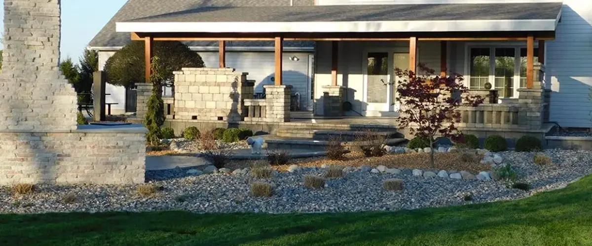 Outdoor living space with fresh, green lawn, stone grill, landscape design with small stones and plants, and a covered patio.