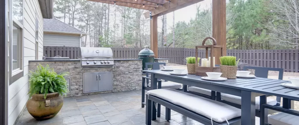 Pretty summer outdoor kitchen with table set and grilling station underneath wooden arbor on stone patio.