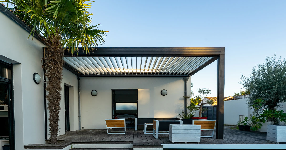 A modern patio with a louvered pergola, wooden deck, minimalist furniture, and a nearby pool. Palm trees and greenery add a tropical vibe