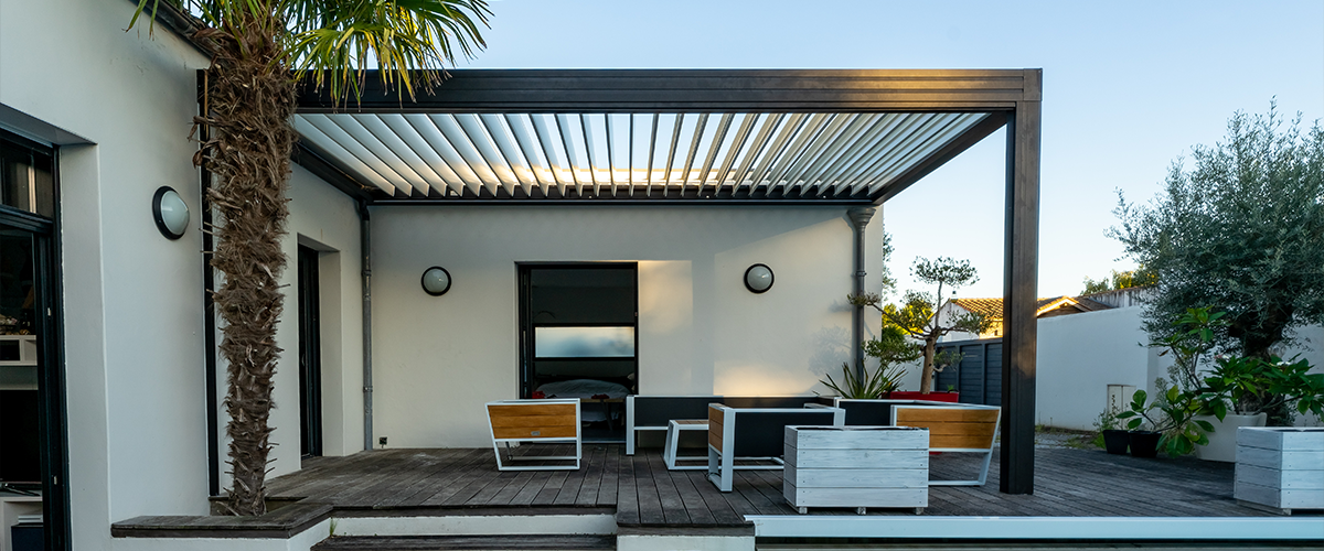 A modern patio with a louvered pergola, wooden deck, minimalist furniture, and a nearby pool. Palm trees and greenery add a tropical vibe