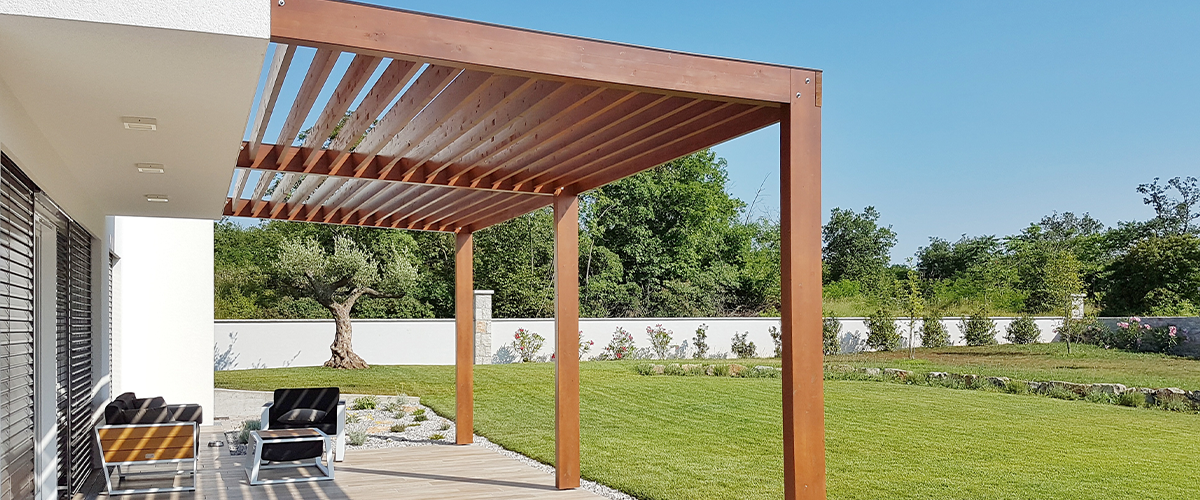 A modern patio with a wooden pergola, sleek outdoor furniture, and a spacious green lawn, enclosed by a white privacy wall and surrounded by trees.